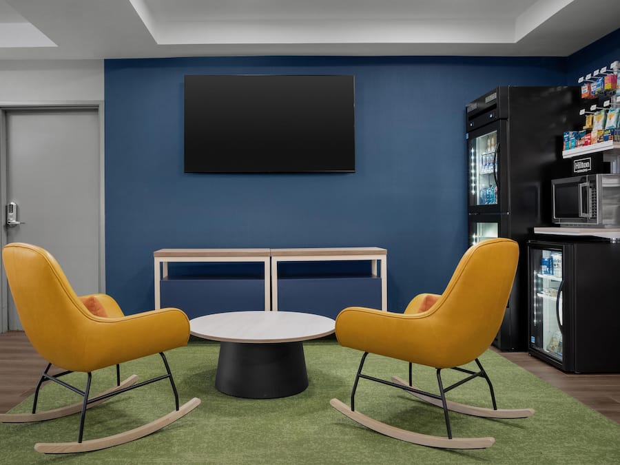 A modern room with a blue wall, a black TV mounted above a wooden table with two yellow rocking chairs, and a minibar with a refrigerator and microwave.