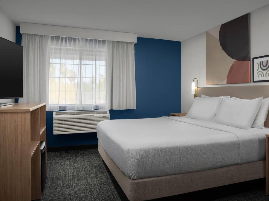 Ein Hotelzimmer mit Bett, Schreibtisch, Fernseher, einem Fenster mit Vorhängen und Bildern an der Wand.