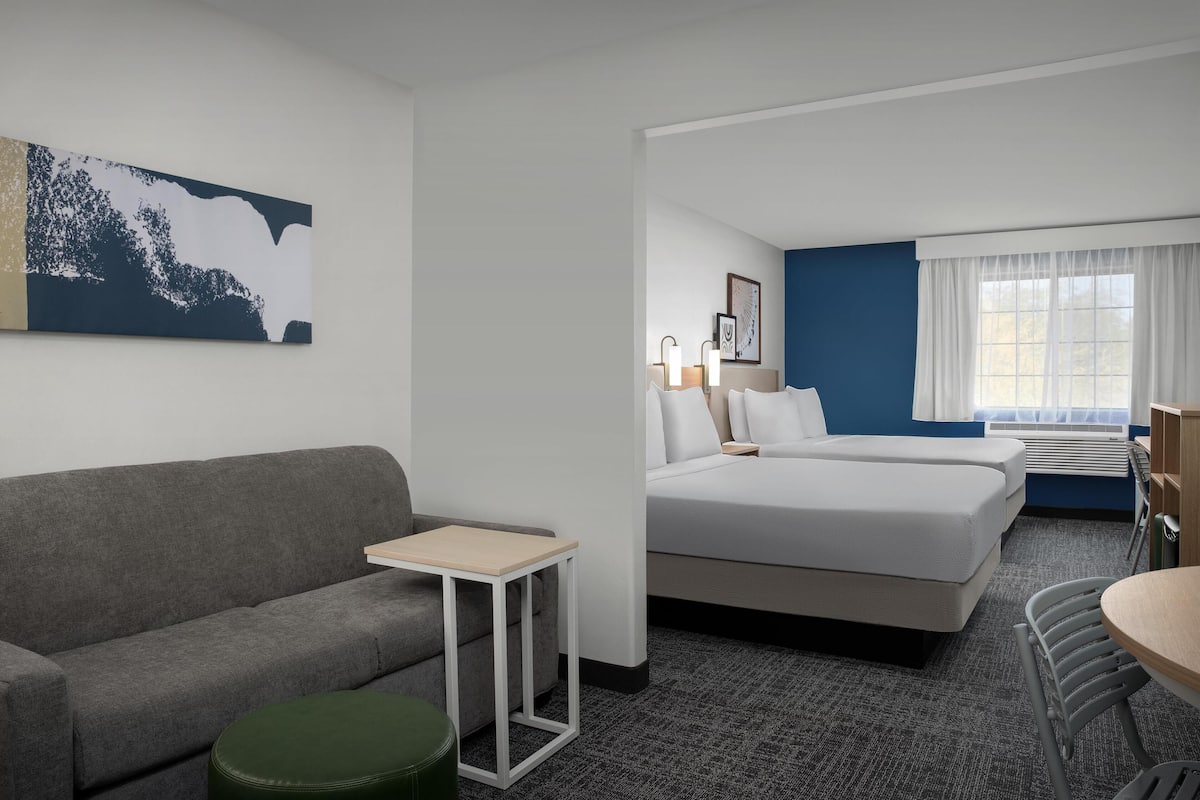 Een hotelkamer met een grijze bank, een klein tafeltje, een groene poef, een bed met witte lakens, een nachtkastje met lampen, een raam met witte gordijnen en een schilderij aan de muur.