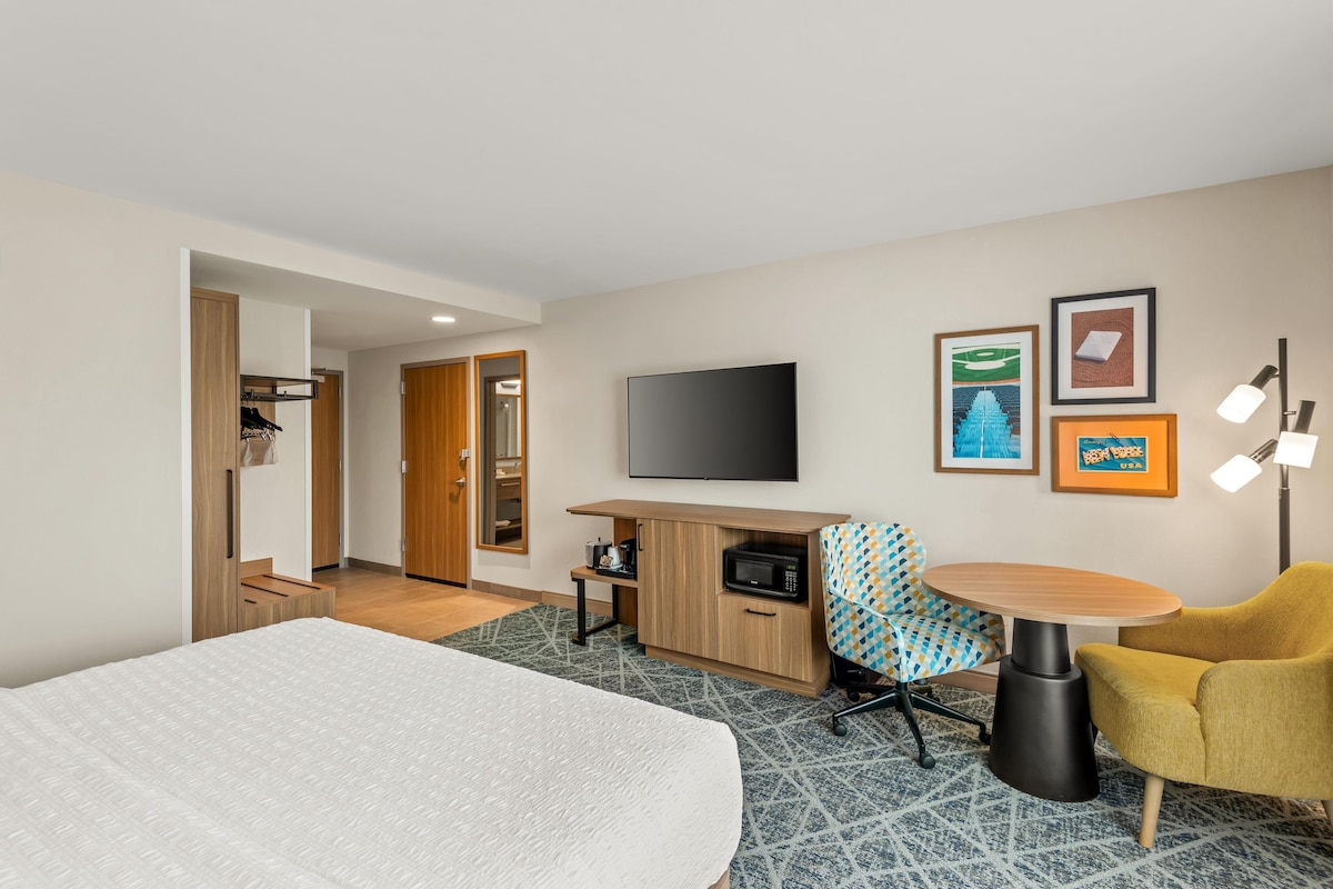 A hotel room with a bed, a desk with a flat-screen TV, a microwave, a chair, and a small table.
