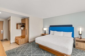 A hotel room with a large bed, a wardrobe, a small desk, and a nightstand with a lamp.