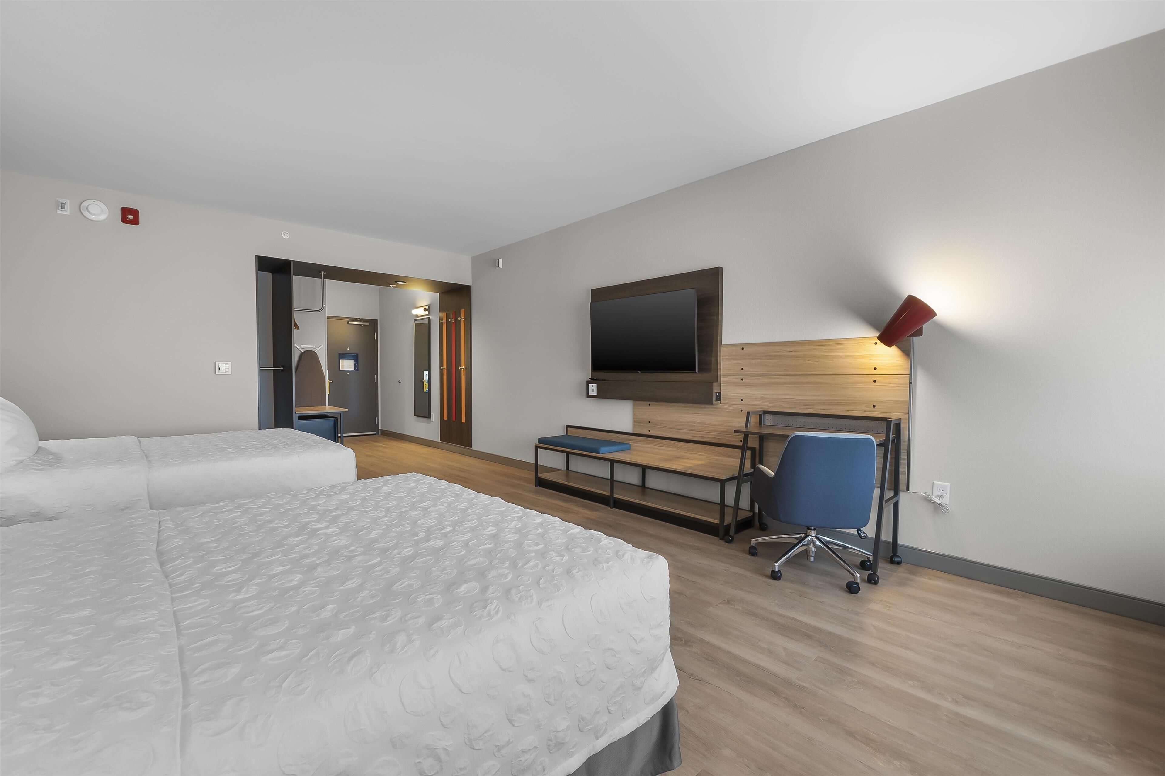 A modern hotel room with two beds, a desk with a chair, a TV mounted on the wall, and a red floor lamp.
