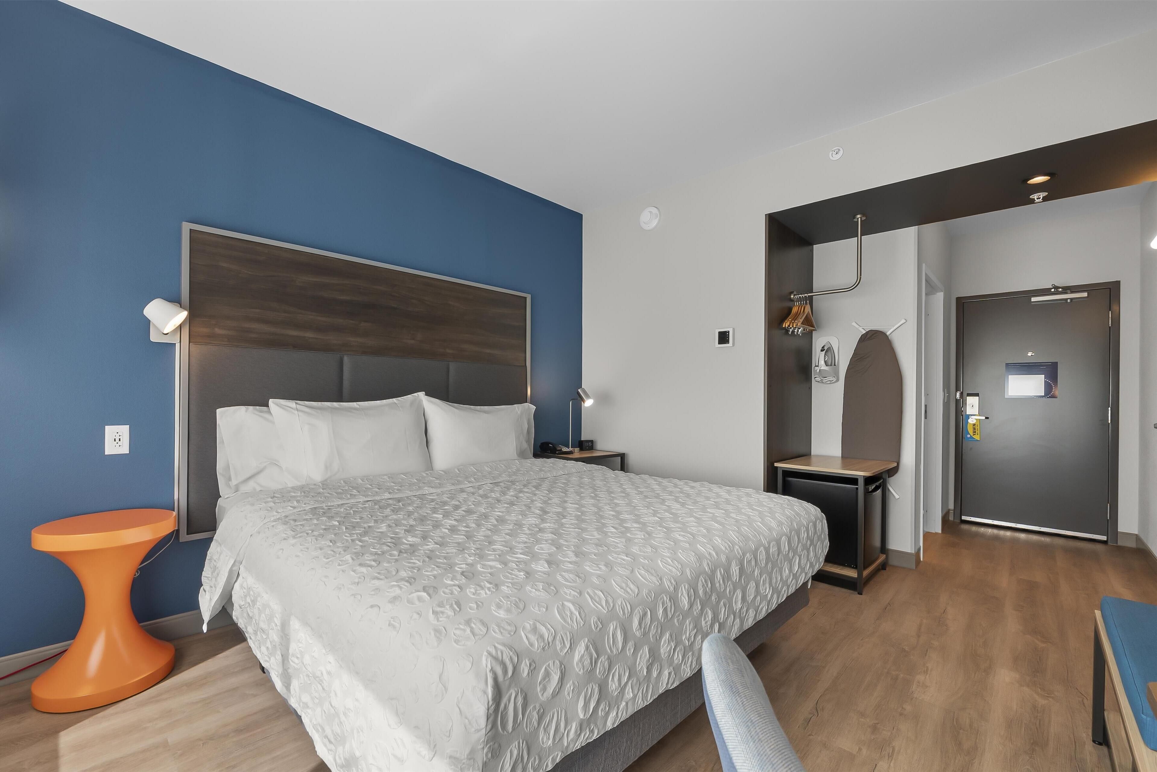 A modern hotel room with a large bed, a small orange side table, a desk, and a chair.