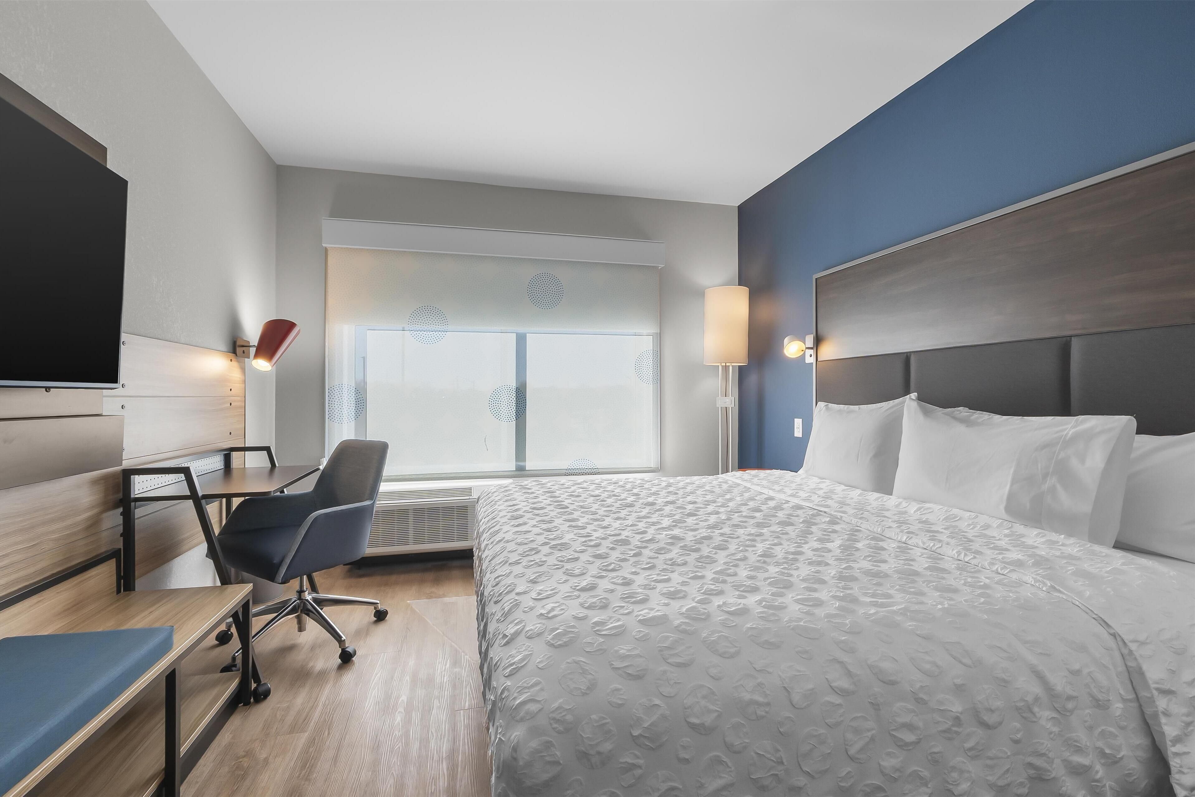 A hotel room with a bed, a desk, a chair, a TV, and a window with blinds.