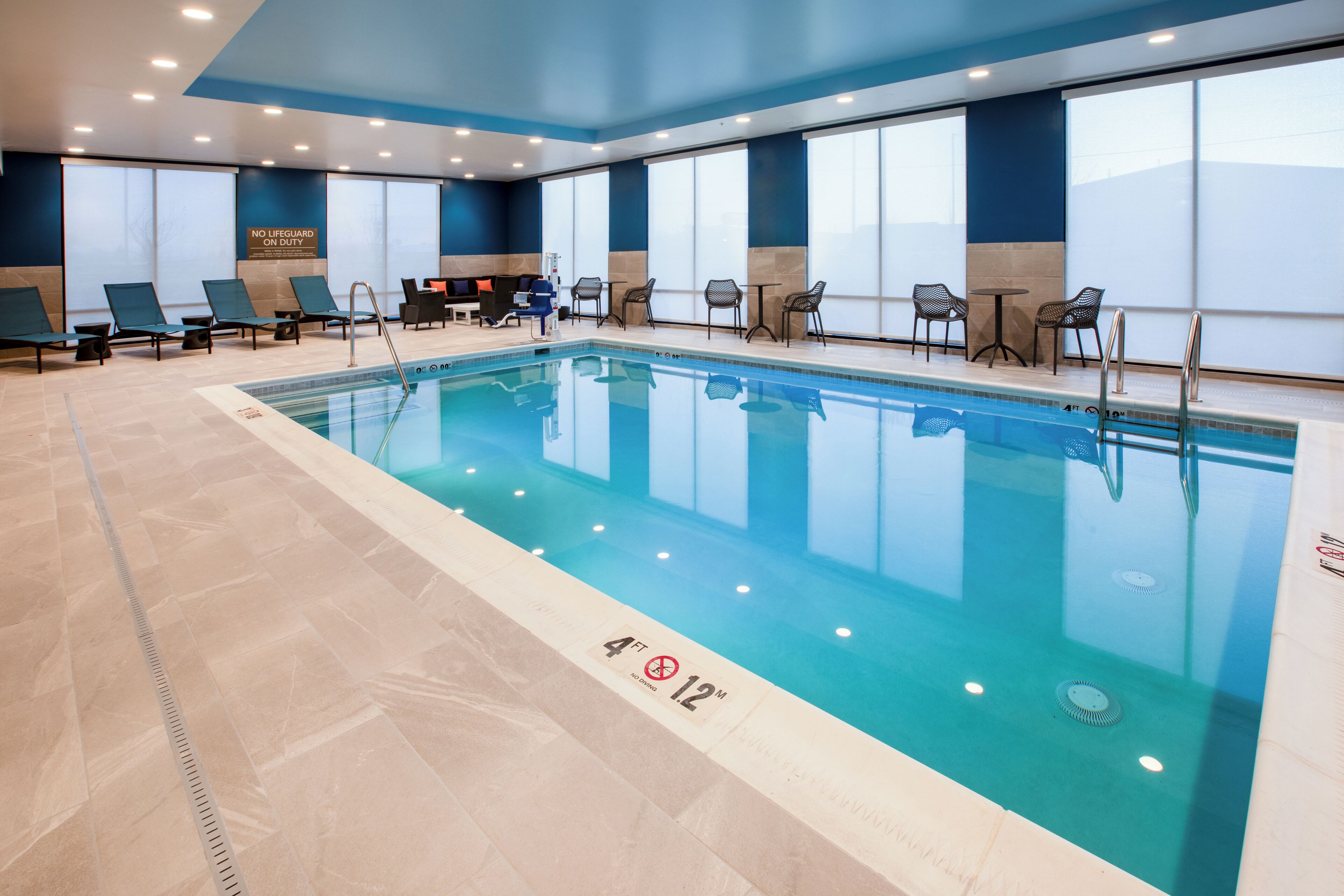 An indoor swimming pool with lounge chairs and tables, a lifeguard sign, and a pool depth marker.