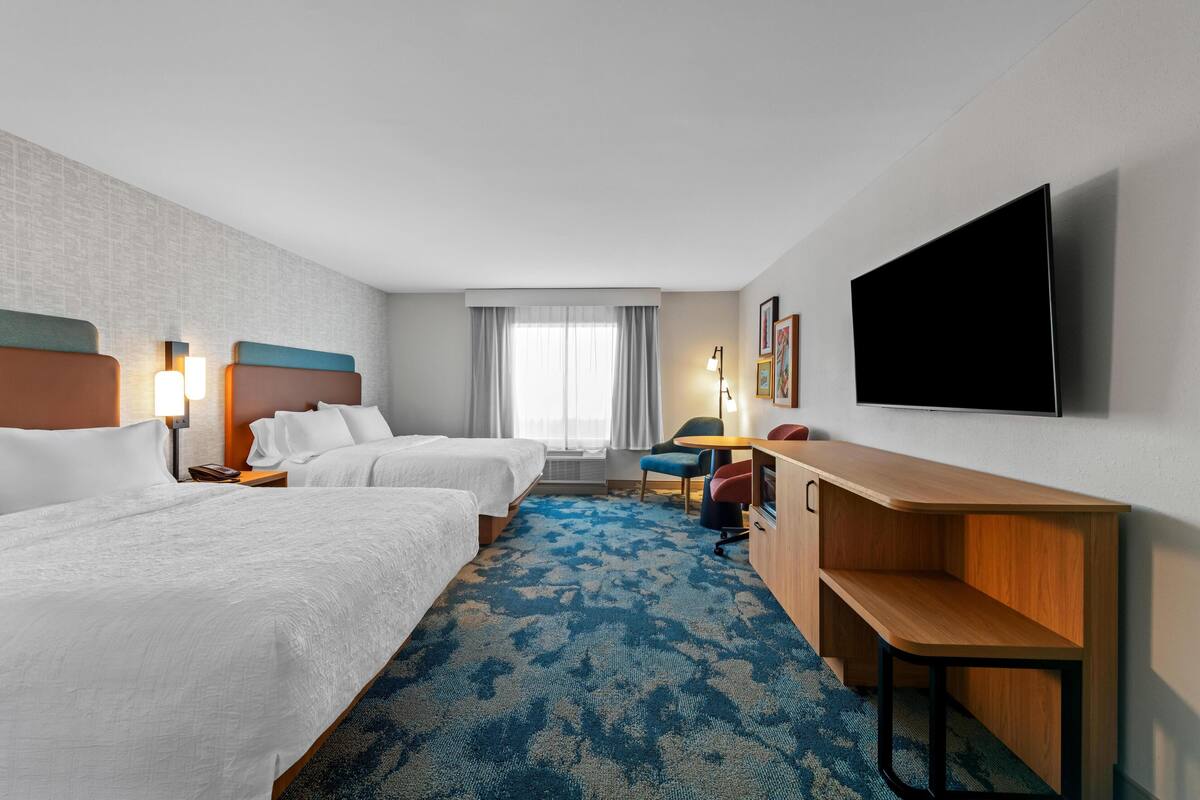 Ein Hotelzimmer mit zwei Betten, einem Schreibtisch, einem Stuhl, einem Flachbildfernseher und einem Fenster mit Vorhängen.