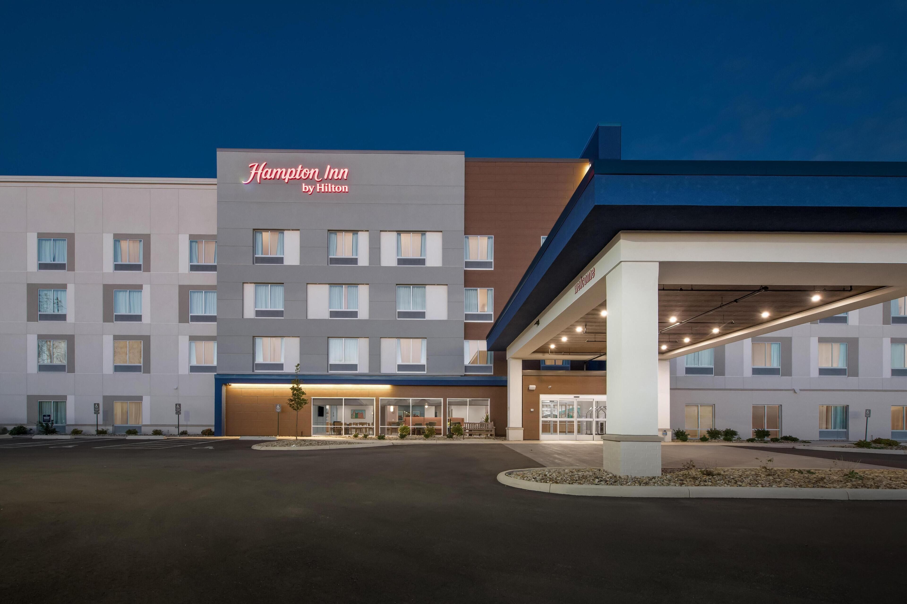 A Hampton Inn by Hilton hotel with a modern facade, multiple floors, and a well-lit entrance.