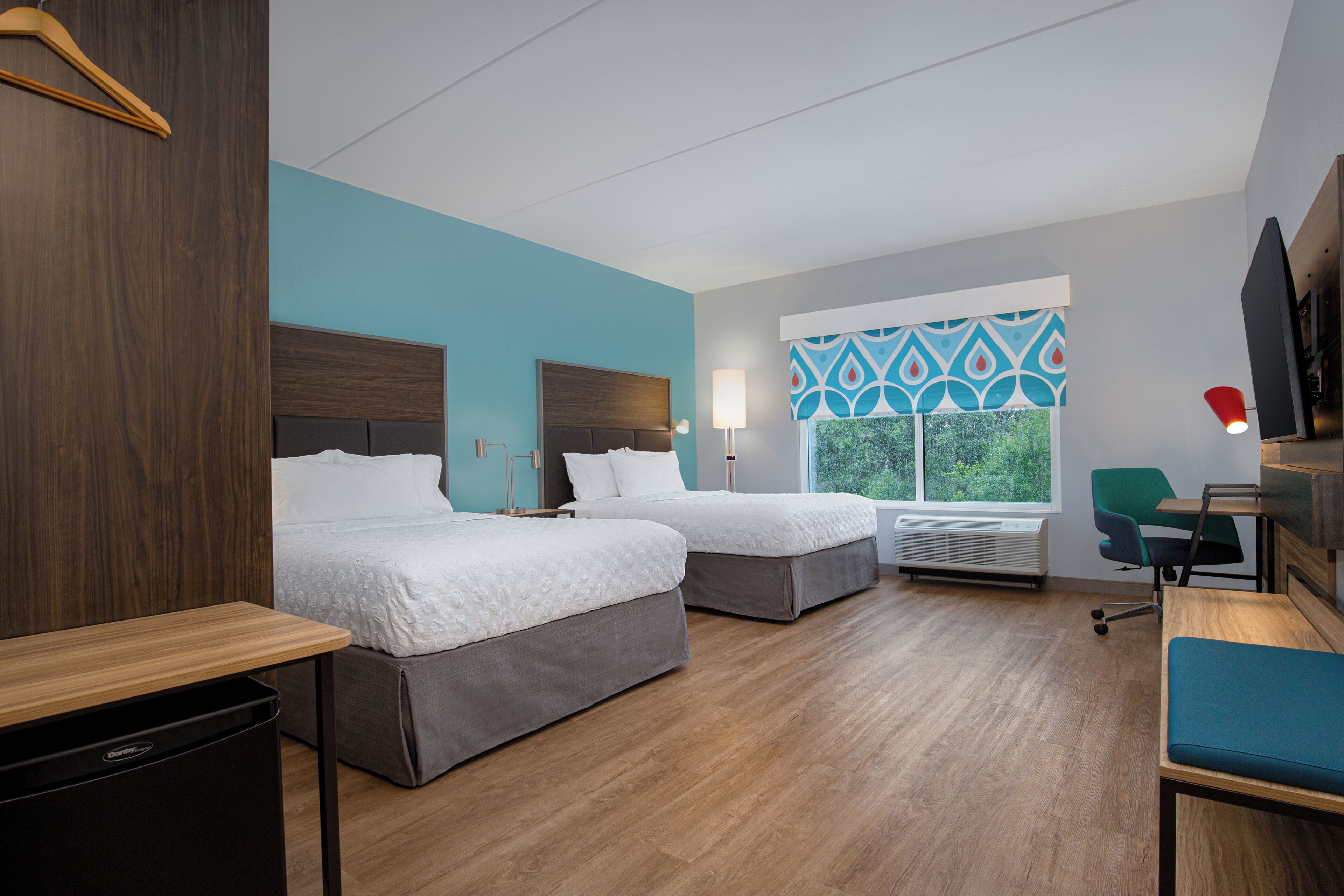 A hotel room with two beds, a desk, a chair, and a window with a view of greenery.