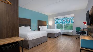 A hotel room with two beds, a desk, a chair, and a window with a view of greenery.