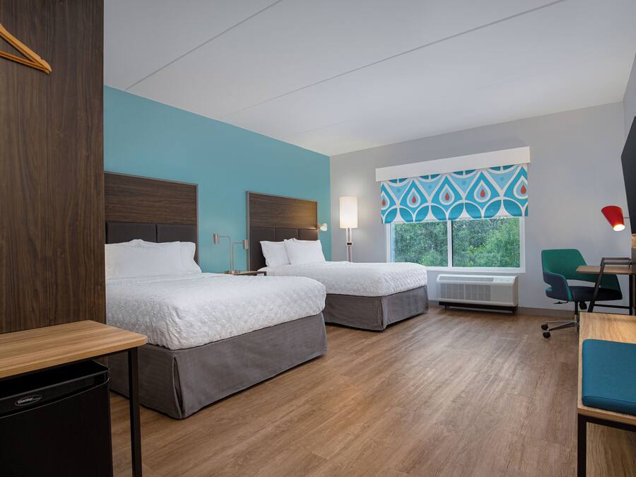 A hotel room with two beds, a desk, a chair, and a window with a view of greenery.