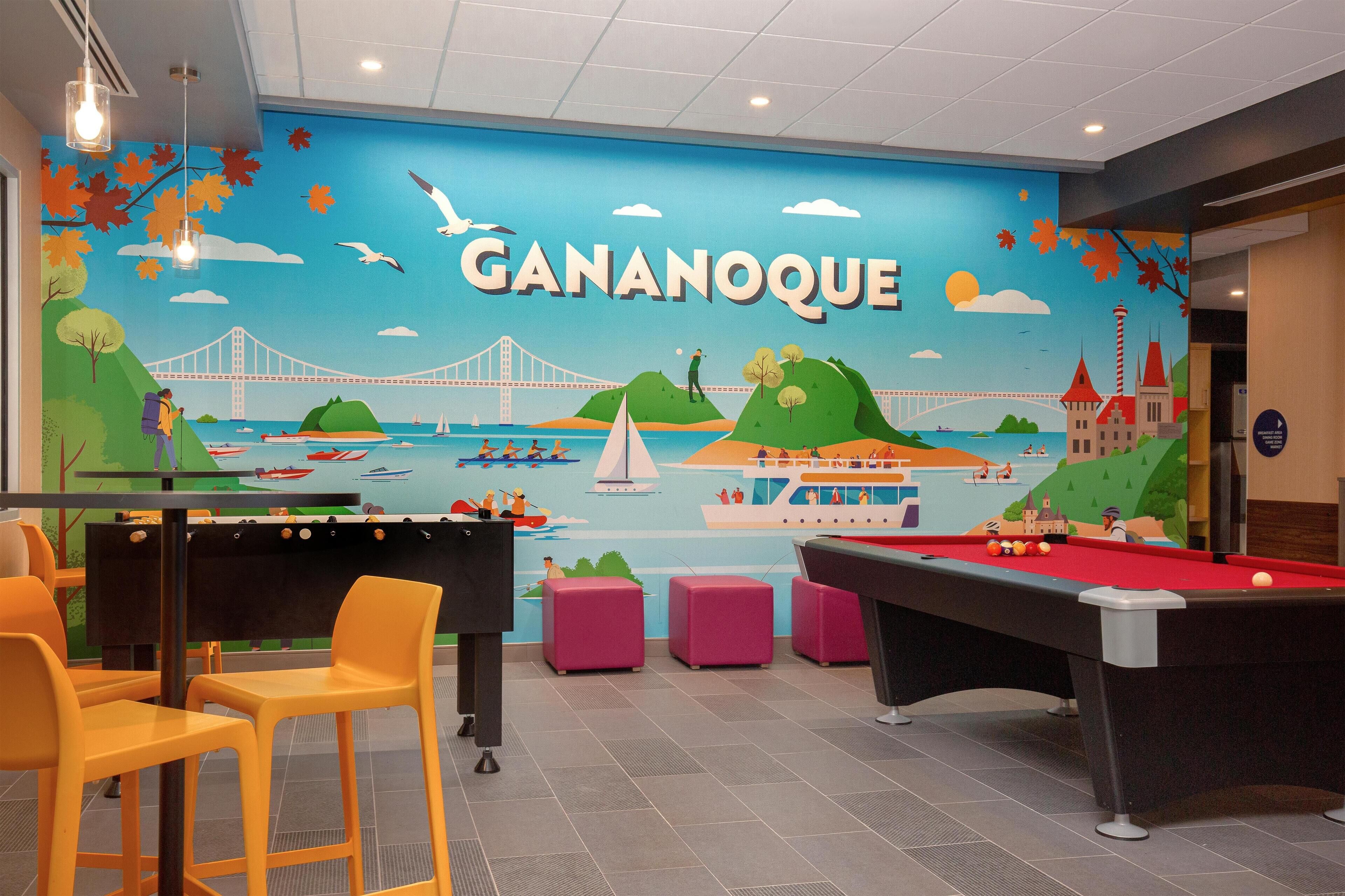 A pool table and chairs in a room with a mural of Gananoque, a bridge, and boats.