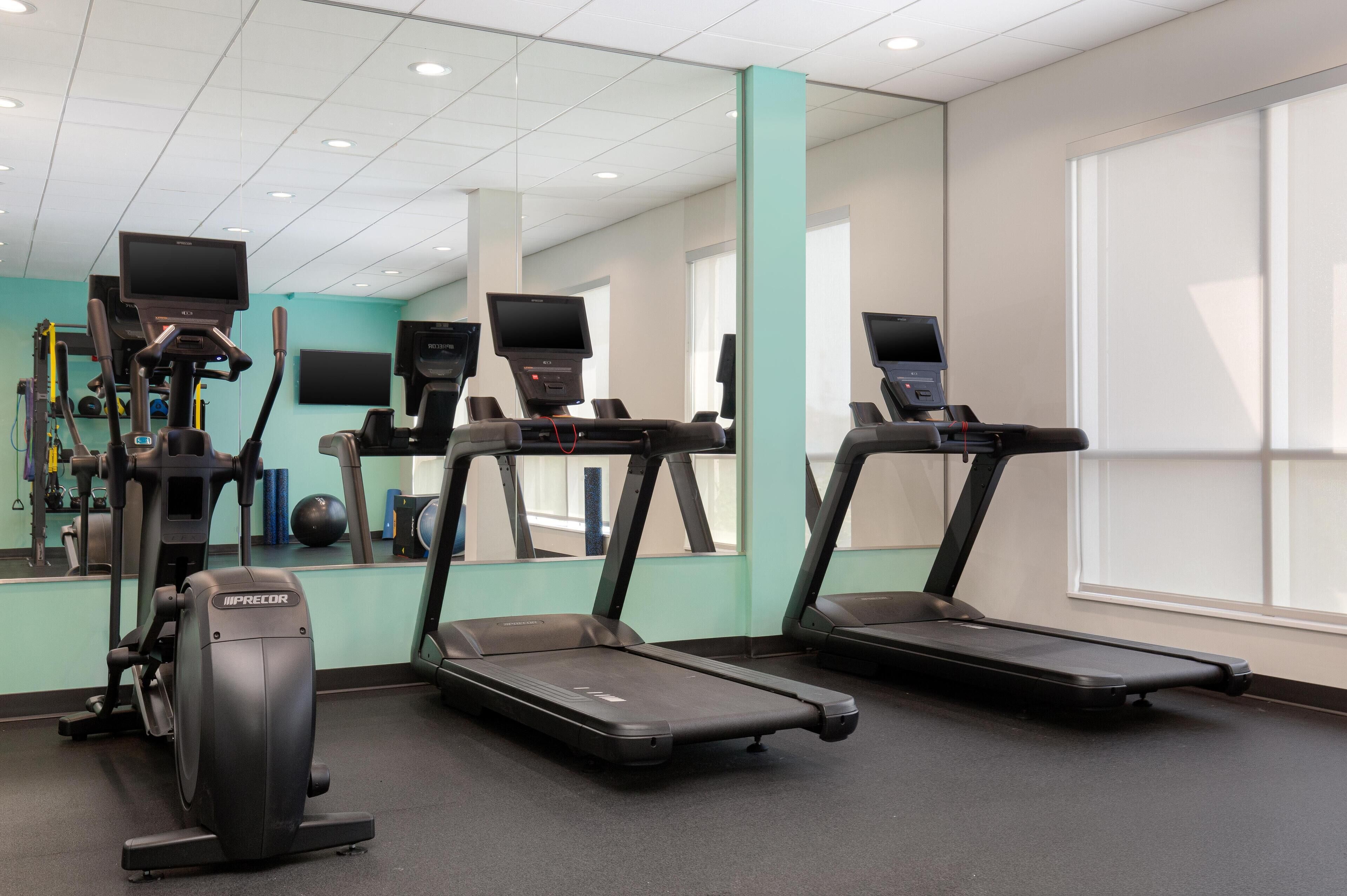 A gym with treadmills, an elliptical, and a stationary bike, with a large mirror and windows.