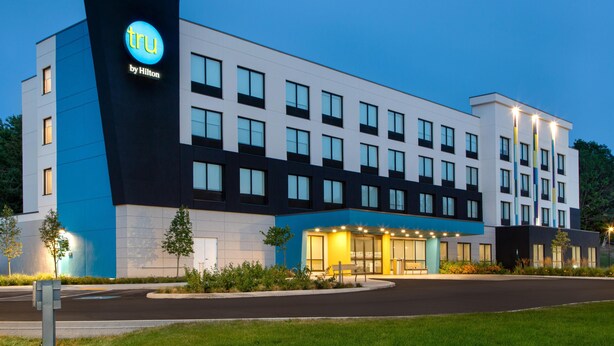 Ein modernes Hotelgebäude mit einem gut sichtbaren 'tru by Hilton'-Schild, zahlreichen Fenstern und einem gut beleuchteten Eingangsbereich.