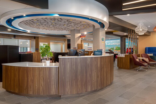 A hotel reception area with a wooden reception desk, a digital display, and a seating area with chairs.