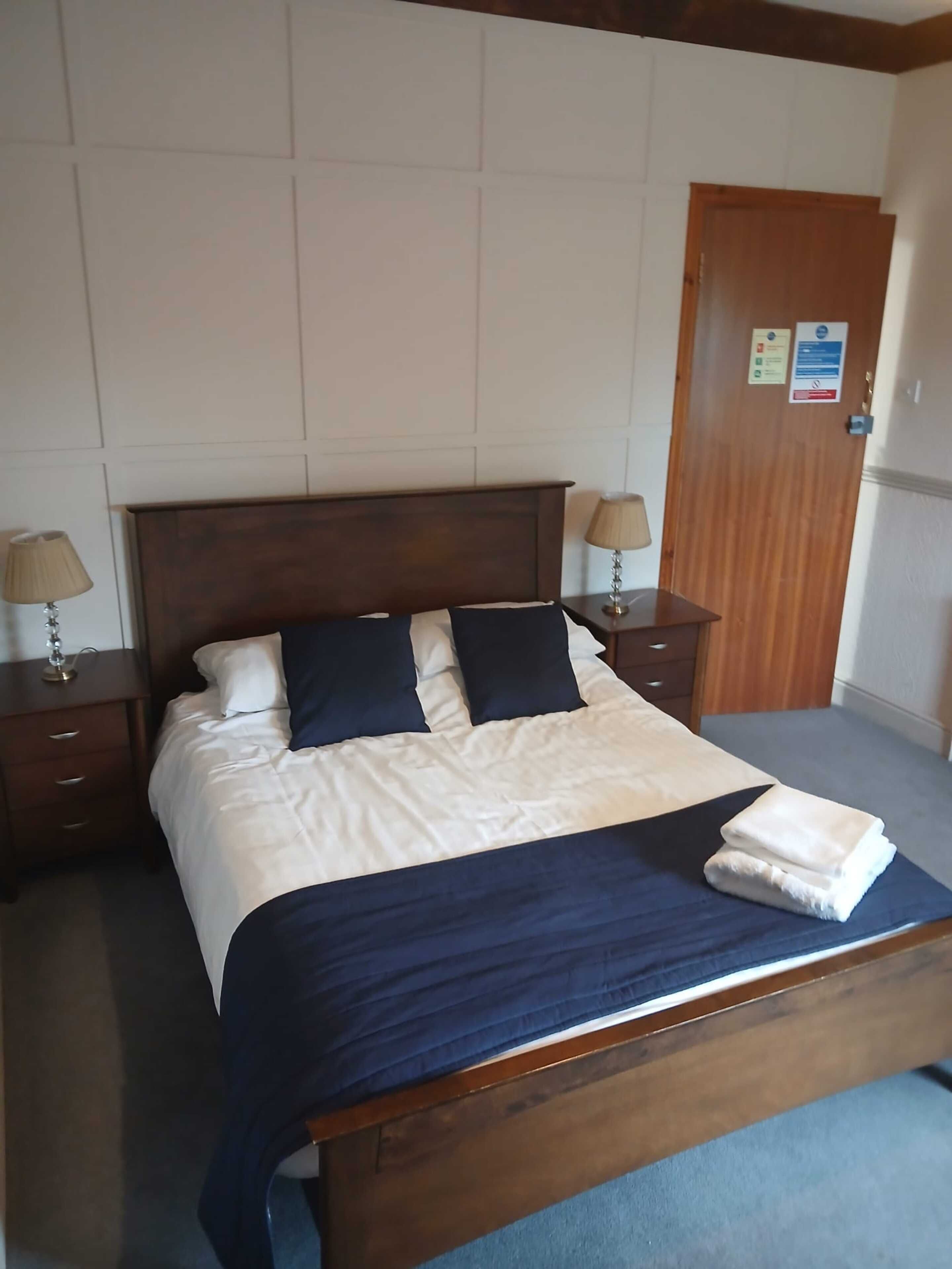 Una cama bien hecha con una colcha y almohadas azul oscuro, cabecera de madera, dos mesitas de noche con lámparas y una puerta de madera con un letrero.