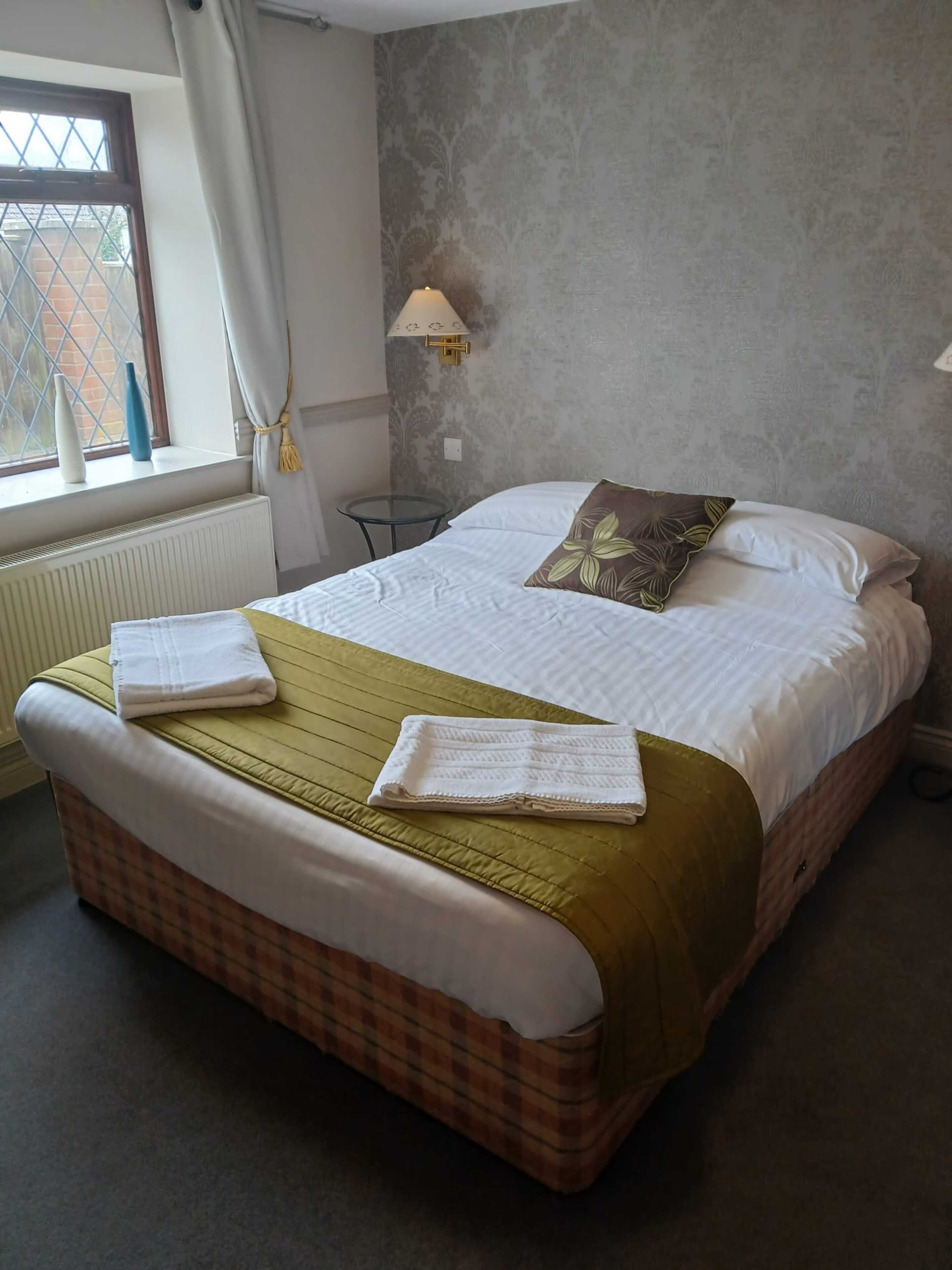 A neatly made bed with a checkered bedspread, two towels, and a single decorative pillow. A small table and a wall-mounted lamp are visible in the room.