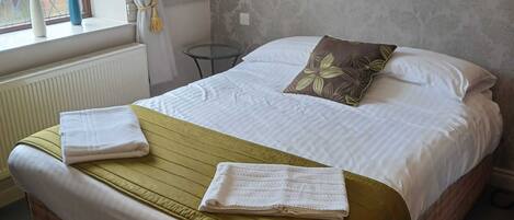 A neatly made bed with a checkered bedspread, two towels, and a single decorative pillow. A small table and a wall-mounted lamp are visible in the room.