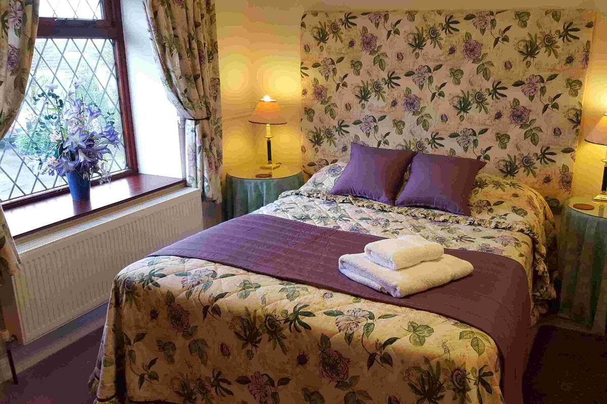 A bedroom with a floral bedspread, two bedside lamps, a bedside telephone, and a window with curtains.