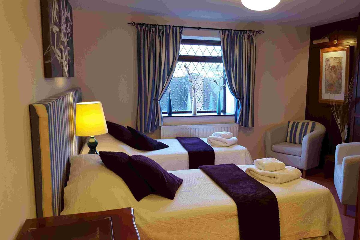 A hotel room with two beds, a chair, a lamp, and a window with curtains.