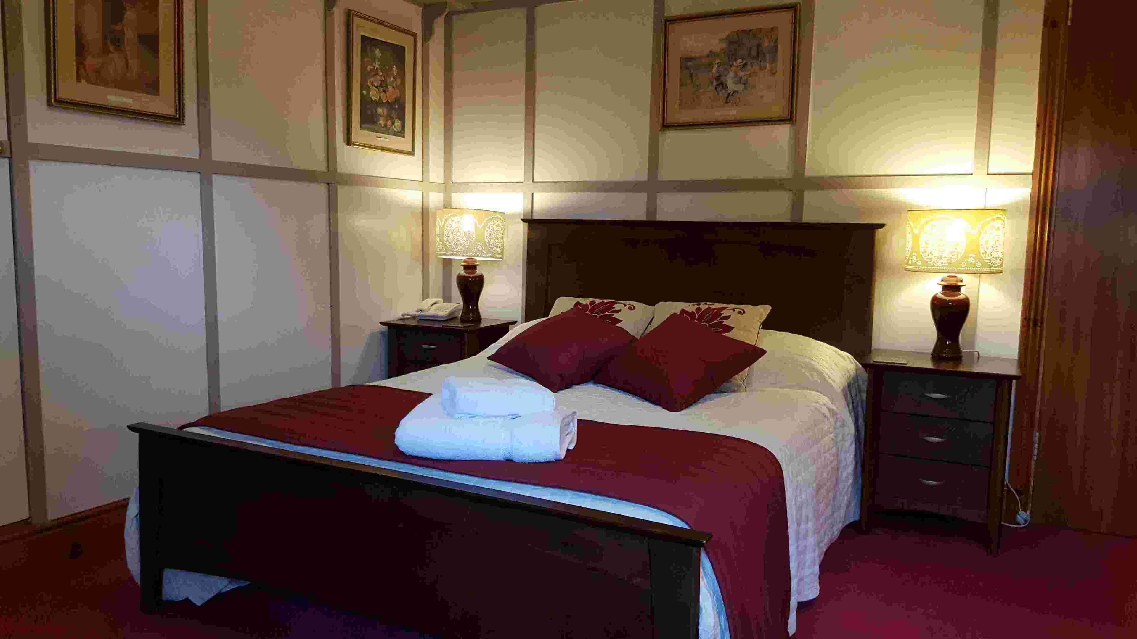 Una cama bien tendida con una colcha roja y almohadas, flanqueada por dos mesitas de noche con lámparas, en una habitación con cuadros en las paredes.