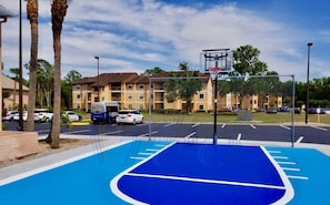 A basketball court with a blue surface, surrounded by a chain-link fence, in front of a multi-story building with balconies.