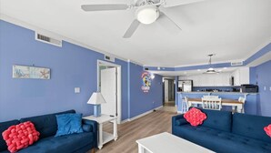 A living room with two blue sofas, a white coffee table, a ceiling fan, and a kitchen area with white cabinets and a bar. - OCbeachparadise, Paradise Off The Boardwalk (Ocean City)