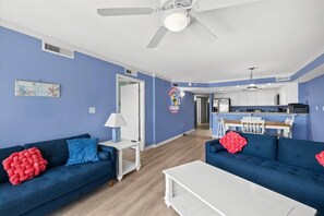 A living room with two blue sofas, a white coffee table, a ceiling fan, and a kitchen area with white cabinets and a bar. - OCbeachparadise, Paradise Off The Boardwalk (Ocean City)