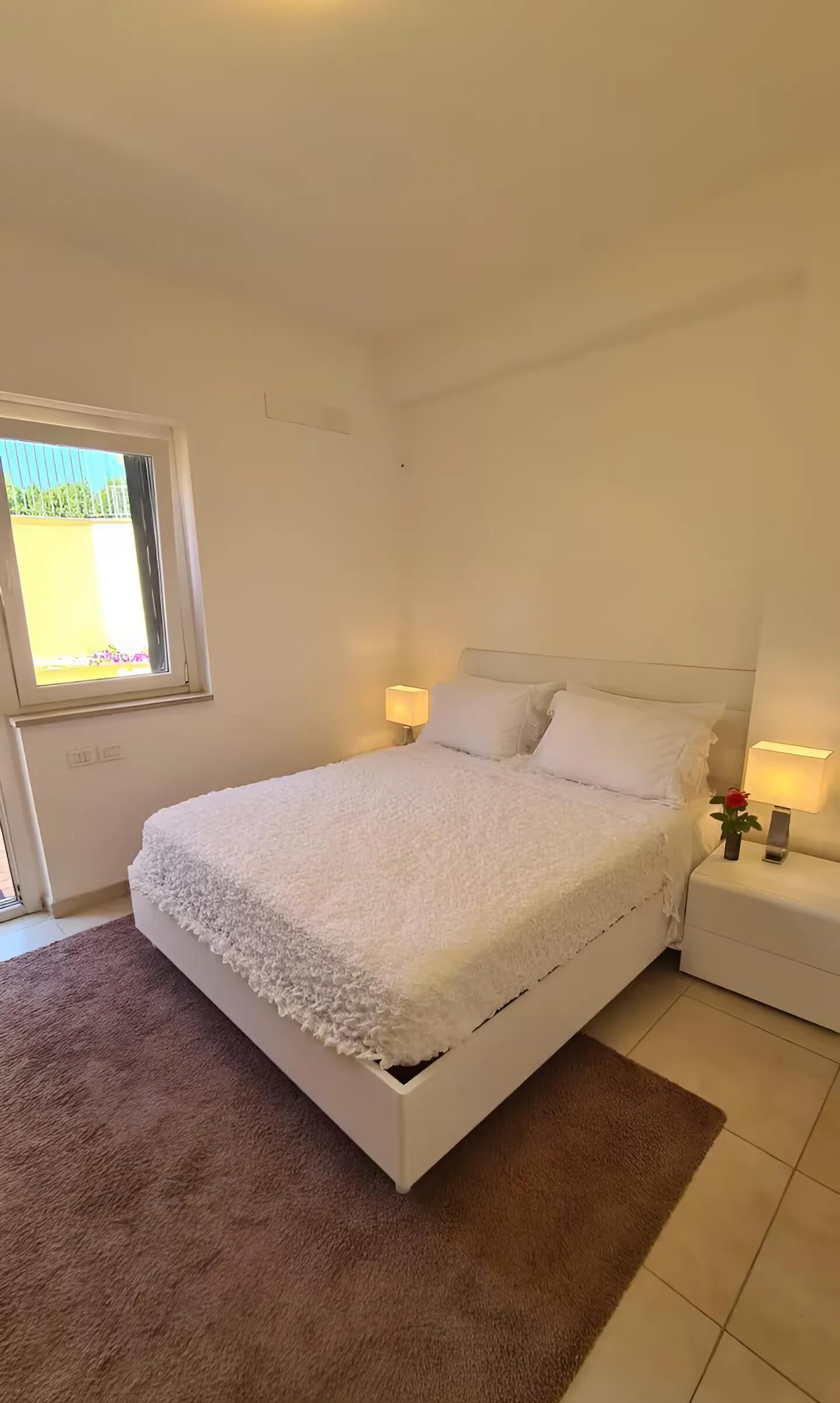 Een slaapkamer met een eenpersoonsbed, twee bedlampjes, een raam en een bruin vloerkleed.