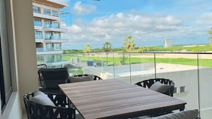 A balcony with a wooden table and wicker chairs, overlooking a residential area with palm trees and a distant building. - Brand new apt w 2 Bedrooms Terrace in Cap Cana (Punta Cana)
