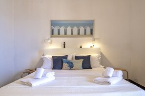 Una cama bien hecha con ropa blanca y toallas, un cuadro encima y lámparas de noche.