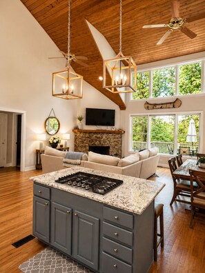 A spacious kitchen with a central island, a stone fireplace, and a dining area with a view of greenery.
