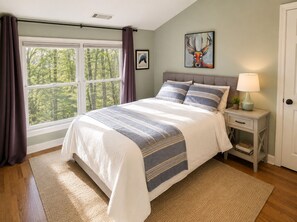 A bedroom with a large bed, a nightstand with a lamp, a framed picture on the wall, and a window with curtains.