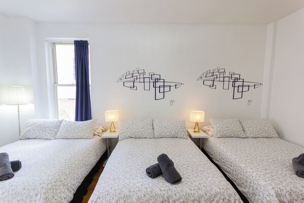 Hotelkamer met twee bedden, elk voorzien van een bloemenprint deken, een hoofdkussenpaar en een opgerolde handdoek. Twee nachtkastjes met lampen, een raam met blauw gordijn, en een kunstwerk boven de bedden.