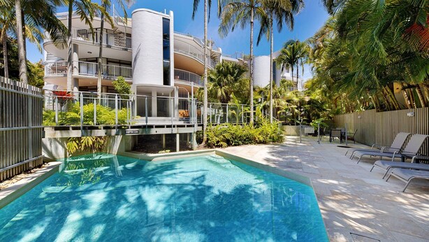 Una piscina con agua cristalina azul, rodeada de palmeras y un edificio con balcones.