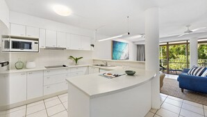 Una cocina moderna con armarios blancos, una isla central, un microondas y un comedor con un sofá azul y vistas al exterior.