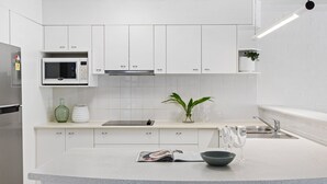 Una cocina moderna con armarios blancos, microondas integrado, refrigerador de acero inoxidable, una isla de vidrio con un tazón y una revista, y una planta en maceta.