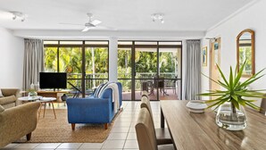 Amplia sala de estar con un sofá azul, una mesa de comedor de madera y un televisor. Amplias ventanas ofrecen vistas a exuberante follaje.