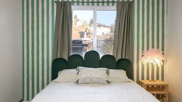 Cama com edredom branco, cabeceira estofada verde, cortinas e abajur com cúpula de penas.