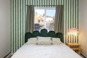 A bed with a white comforter and a green upholstered headboard, curtains, and a bedside lamp with a feathered shade.