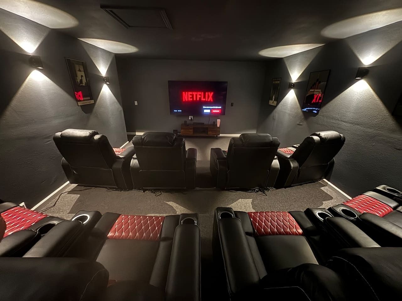 Sala de cinema com uma TV de tela grande exibindo o logotipo da Netflix, várias fileiras de poltronas de couro reclináveis pretas e alto-falantes fixados na parede.