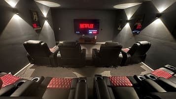 Sala de cinema com uma TV de tela grande exibindo o logotipo da Netflix, várias fileiras de poltronas de couro reclináveis pretas e alto-falantes fixados na parede.