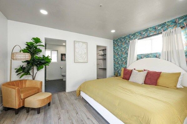 Une chambre à coucher avec un grand lit, une chaise, une plante et une salle de bain visible à travers une porte ouverte.