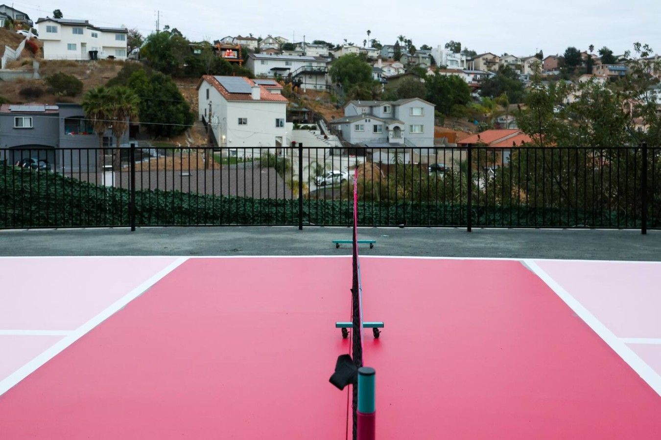 En udendørs tennisbane med et net i midten, omgivet af et sort metalhegn, og boligbygninger i baggrunden.