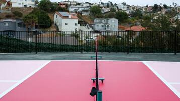 Un campo da tennis all'aperto con una rete al centro, circondato da una recinzione in metallo nero, e palazzi residenziali sullo sfondo.