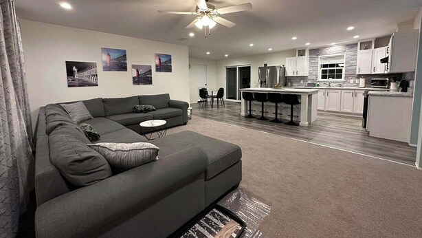 Ein modernes Wohnzimmer mit einem grauen Ecksofa, einem Deckenventilator und einer Küchenzeile mit weißen Schränken und einer Marmor-Spritzschutzwand.