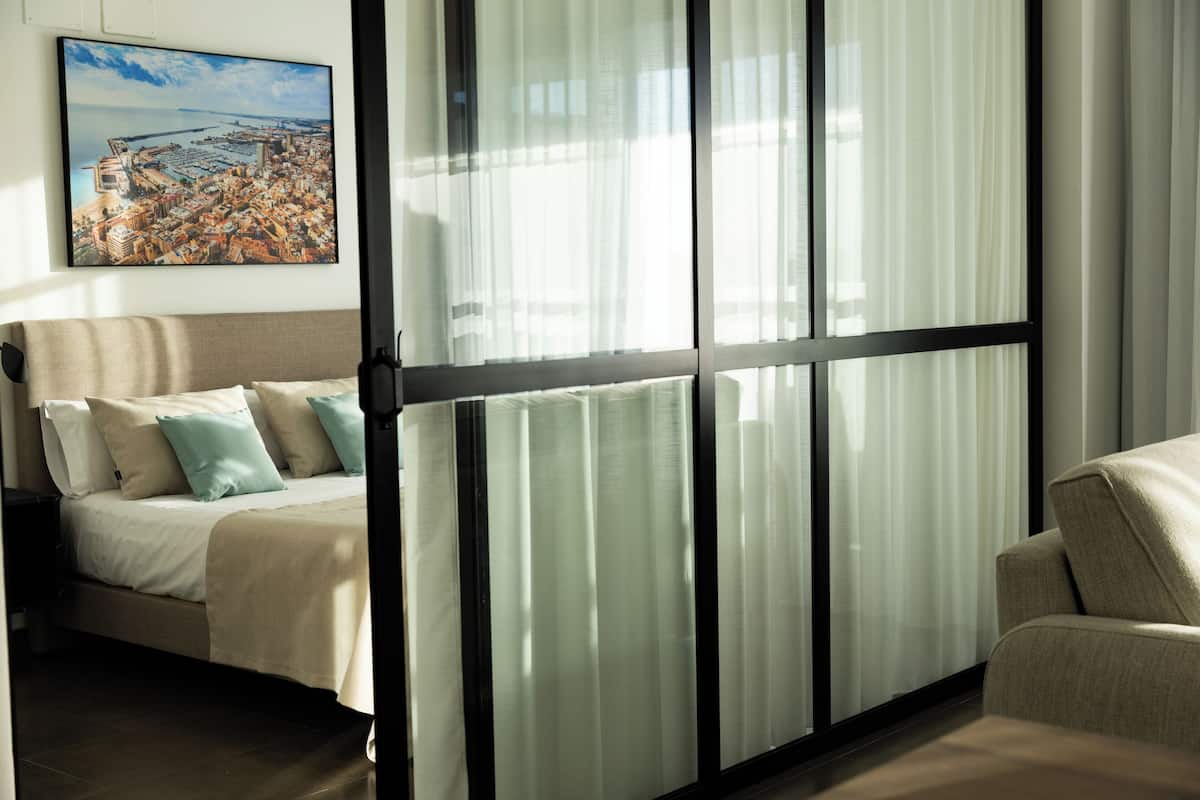 Une chambre d’hôtel moderne dotée d’une grande fenêtre, d’un lit avec une literie beige et des coussins turquoise, et offrant une vue sur le paysage urbain.