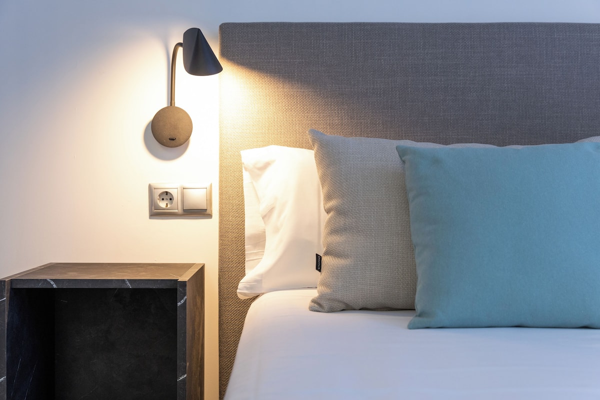 Un lit bien fait, avec des oreillers blancs et bleu clair, une lampe fixée au mur et une prise électrique sur un mur à texture.