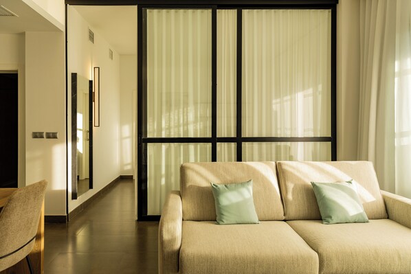 Una sala de estar moderna con un sofá color beige, dos almohadas, una silla de madera y un ventanal con cortinas translúcidas.