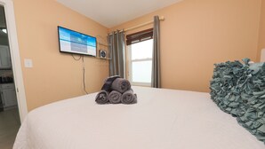 Ein Schlafzimmer mit einem Bett, einem an der Wand befestigten Fernseher, einem Fenster mit Vorhängen und Blick ins Freie.