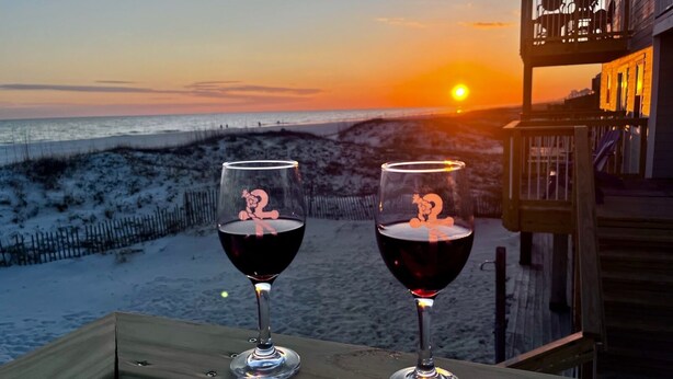 Zwei Weingläser mit Zeichentrickfiguren auf einem hölzernen Deck mit Blick auf einen Strand bei Sonnenuntergang.